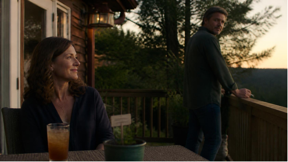 Woman sitting at a deck table with an iced tea at sunset while a man stands at the railing behind her, holding a cigarette and looking back at her