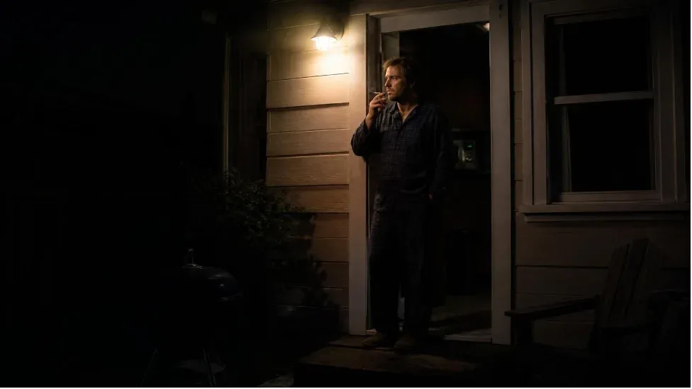 A man in pajamas standing in a lit doorway at night, smoking his last cigarette before bed